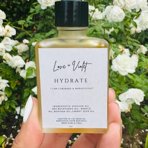 Puede incluir: Una botella de vidrio de aceite facial Love Violet Hydrate con un gotero negro. La etiqueta es blanca con texto negro que dice "Love Violet Hydrate" y "Soy luminosa y magnífica". Los ingredientes se enumeran como: Aceite de aguacate, aceite de espino amarillo, aceite de marula, aceite de moringa, aceite de semilla de zanahoria. La botella se elabora en Los Ángeles y nutre el cuerpo, la mente y el alma. La botella es de 1 oz.