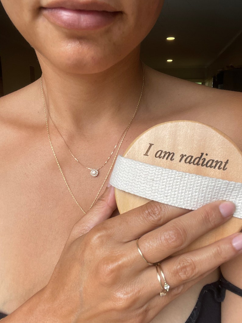 Puede incluir: Un cepillo corporal de madera con una correa blanca. El cepillo tiene las palabras "Soy radiante" grabadas.
