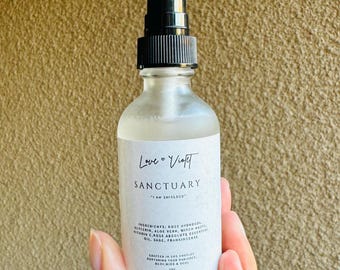 May include: A clear glass bottle with a black spray top, labeled "Love Vioft Sanctuary" and "I am shielded". The bottle contains a clear liquid and has a list of ingredients printed on the label.