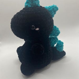May include: A handmade black crochet dinosaur with teal accents. The plush toy has a rounded body, small legs, and a row of teal spikes down its back. It has small, round eyes and is sitting on a white surface.