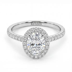 Może przedstawiać: Diamentowy pierścionek zaręczynowy z oprawą halo. Pierścionek ma duży diament o owalnym szlifie otoczony mniejszymi diamentami. Opaska jest również inkrustowana diamentami. Na opasce wygrawerowano napis "RITZIN".