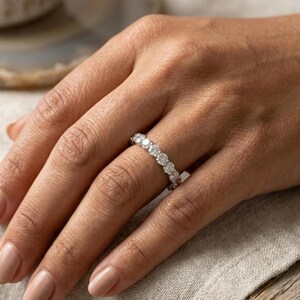 Pode incluir: Um anel de eternidade prateado com uma série de diamantes redondos. O anel é exibido em um dedo, com a mão repousando sobre um tecido de cor neutra. Os diamantes brilham.