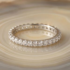 Puede incluir: Un anillo de eternidad plateado con una fila continua de piedras preciosas redondas y claras. El anillo está colocado sobre una superficie clara y arremolinada, creando un contraste que resalta el brillo del anillo. El diseño del anillo es simple y elegante.