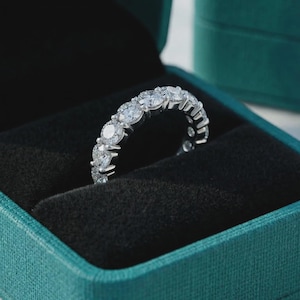 Könnte beinhalten: Ein silberner Eternity-Ring mit mehreren runden, klaren Edelsteinen. Der Ring ist in einem silbernen Band gefasst und wird in einer türkisfarbenen Schmuckschatulle mit schwarzem Samtinterieur präsentiert. Ein klassisches Schmuckstück.