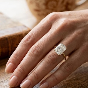 Puede incluir: Un anillo de oro con un gran diamante rectangular de talla radiante. El diamante está engastado en una montura de cuatro garras. El anillo está en una mano con uñas cuidadas. El fondo está borroso.
