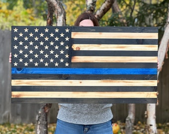 Puede incluir: Una bandera americana de madera con una línea azul en el medio. La bandera está teñida de un color madera oscuro con acentos de madera natural. La bandera está siendo sostenida por una persona.
