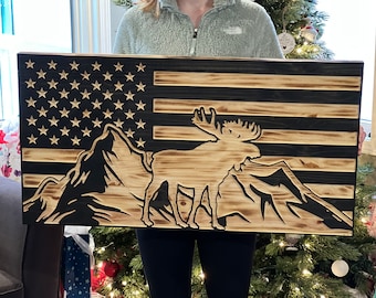 Puede incluir: Una bandera americana de madera con un diseño de silueta de alce y montaña. La bandera está teñida de un color marrón oscuro con un acabado de veta de madera natural.