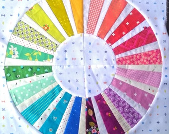 Puede incluir: Un cuadrado de tela blanca con un diseño de patchwork colorido en forma de sol. La tela está compuesta por diferentes patrones y colores, incluyendo rojo, naranja, amarillo, verde, azul y morado.