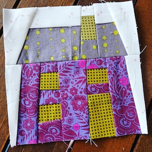 Puede incluir: Un bloque de patchwork colorido con diseños geométricos y florales. El bloque combina telas moradas, amarillas y grises con detalles blancos. El diseño incluye cuadrados y rectángulos, creando un efecto de patchwork.