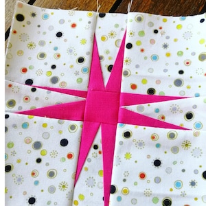 May include: A pink and white patchwork quilt block with a star design. The star is made of pink fabric and the background is white fabric with a pattern of small circles in various colours.