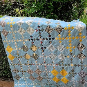 May include: A handmade quilt with a light blue background and a patchwork design. The quilt features squares in various patterns, including polka dots, floral prints, and solid colors of yellow, brown, and white. The quilt is draped over a surface.