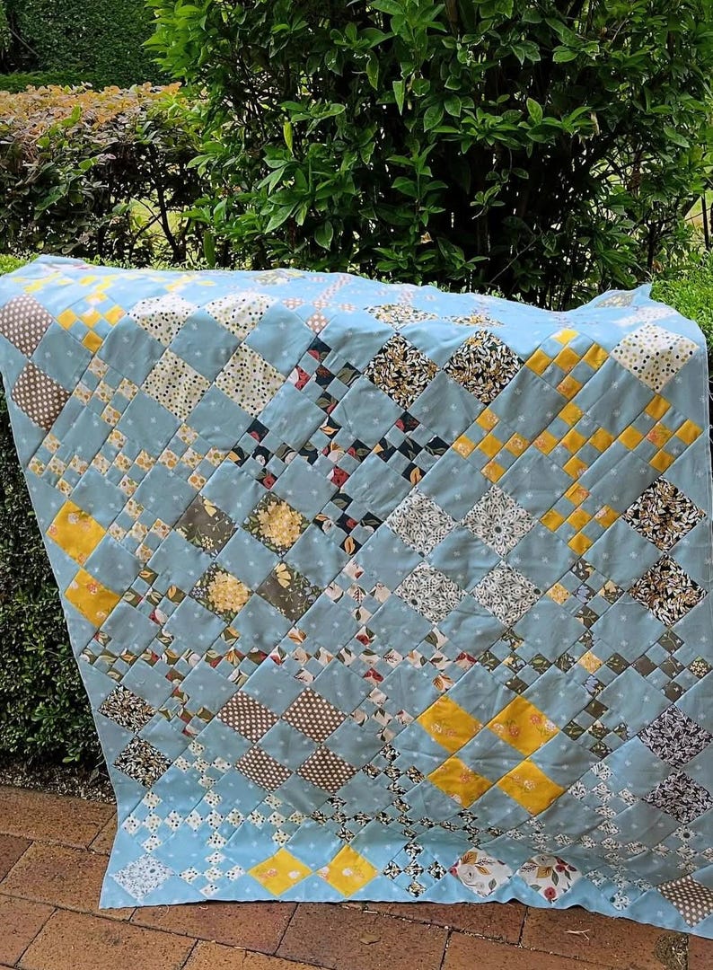 May include: A handmade quilt with a light blue background and a patchwork design. The quilt features squares in various patterns, including polka dots, floral prints, and solid colors of yellow, brown, and white. The quilt is draped over a surface.