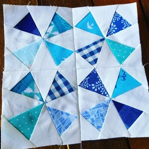 May include: A white quilt block with a patchwork design featuring blue, teal, and white triangles. The triangles are arranged in a repeating pattern, creating a geometric design.