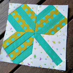 May include: A square fabric quilt block with a gift-like design. The block features teal and yellow patterned fabric, with teal stripes and yellow fabric with blue polka dots. The background is white with small, colorful floral and crescent moon patterns.
