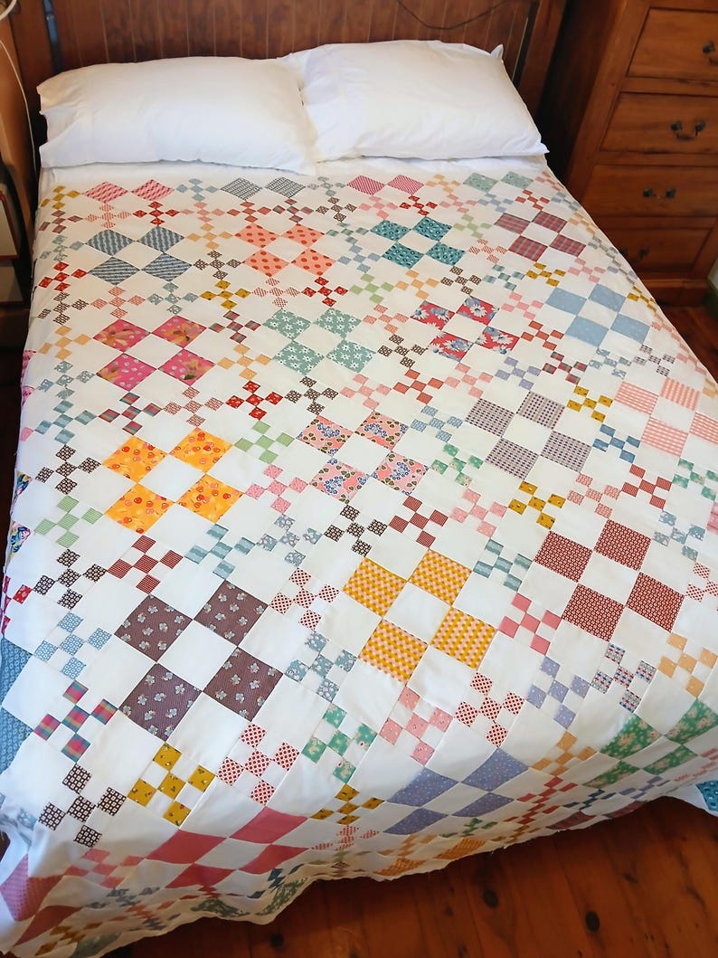 May include: A handmade quilt with a white background and a colorful patchwork design. The quilt features a repeating pattern of squares in various colors, including red, blue, yellow, and pink. The quilt is on a bed with white pillows.