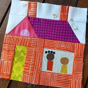 May include: A colorful patchwork quilt block featuring a house with a pink roof, a purple and pink patterned front, and two figures standing in front of it. The house is surrounded by orange and white brick patterns.