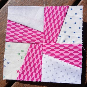 May include: A square quilt block with a pinwheel design. The block features various fabrics in white, pink, and patterned pink. The patterns include polka dots, geometric shapes, and floral motifs. The block is approximately 6 inches square.