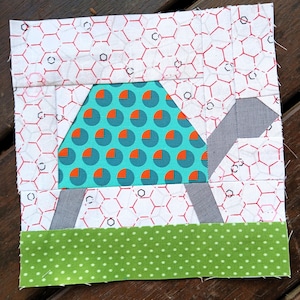 May include: A colorful fabric block featuring a turtle design. The turtle's shell is teal with orange circles, set against a white background with a red honeycomb pattern. Gray legs and a green dotted border complete the design.