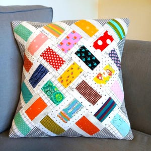 May include: A square throw pillow featuring a patchwork design of rectangular fabric strips in various colors and patterns, including polka dots, florals, stripes, and solid hues, arranged in a diagonal grid on a white background with black grid lines.
