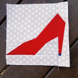 May include: A fabric square featuring a red high-heeled shoe design. The shoe is set against a light gray background with white polka dots. The heel has a small blue accent. This textile art piece is likely for quilting or crafting.