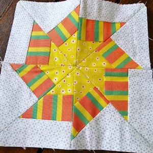 Puede incluir: Un bloque de colcha de retazos con un diseño de estrella. La estrella está hecha de tela amarilla, naranja y verde con rayas. El fondo es una tela gris claro con un pequeño patrón de puntos blancos.