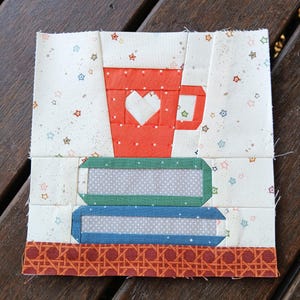 May include: A fabric patchwork block featuring a red mug with a white heart, atop two books. The books are green and blue with gray polka dots. The background is white with star and dot patterns, and a brown patterned border.