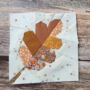Peut inclure: Un bloc de courtepointe en tissu carré présentant une forme de feuille centrale assemblée à partir de divers tissus à motifs marron, orange et jaune moutarde, sur un fond crème clair décoré de petits motifs d'étoiles multicolores.