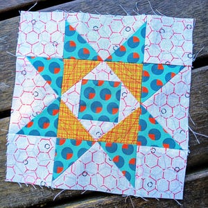 Op de afbeelding: Een vierkant quiltblok met een steruitbarstingontwerp. Het blok heeft turquoise stof met oranje stippen, gele stof met oranje lijnen en witte stof met een rood honingraatpatroon. Het blok ligt op een houten oppervlak.