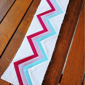 May include: A white fabric strip with a chevron pattern made from turquoise, lavender, and burgundy fabric strips.