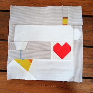 Puede incluir: Un bloque de patchwork hecho a mano con un corazón rojo, rectángulos blancos y tela a rayas grises. También hay toques de amarillo y azul. El bloque es cuadrado y parece estar hecho de retales de tela.