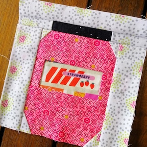 Może przedstawiać: Kwadratowy kawałek materiału w kolorze różowym i białym z czerwoną i białą paskowaną etykietą z napisem "STRAWBERRY" i trzema czerwonymi truskawkami. Kawałek materiału jest blokiem patchworkowym z wzorem różowych i białych kółek.