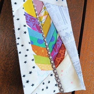 May include: A colorful fabric feather quilt block. The feather design features various patterned fabrics in yellow, purple, blue, orange, and green. The background is white with black star and blue polka dot patterns. The feather is outlined with a brown dotted fabric.