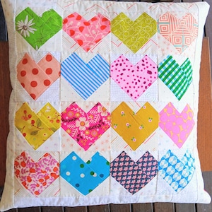 May include: A square throw pillow featuring a patchwork design of sixteen heart-shaped fabric blocks. Each heart displays a unique pattern, including floral, polka dot, striped, and geometric prints in vibrant colors like green, pink, blue, and yellow.