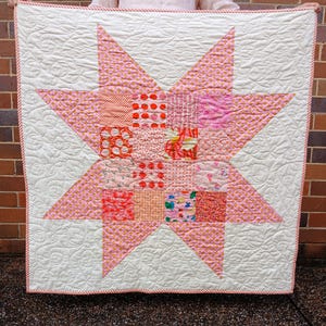 May include: A handmade quilt featuring a large pink star design on a white background. The star is composed of various patterned pink squares. The quilt has a red and white striped border and intricate quilted patterns.