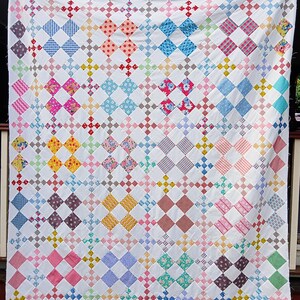 May include: A handmade quilt with a white background and a repeating pattern of colorful diamond shapes. The diamonds are filled with various patterns and colors, including blue, pink, yellow, and red. The quilt is displayed outdoors.