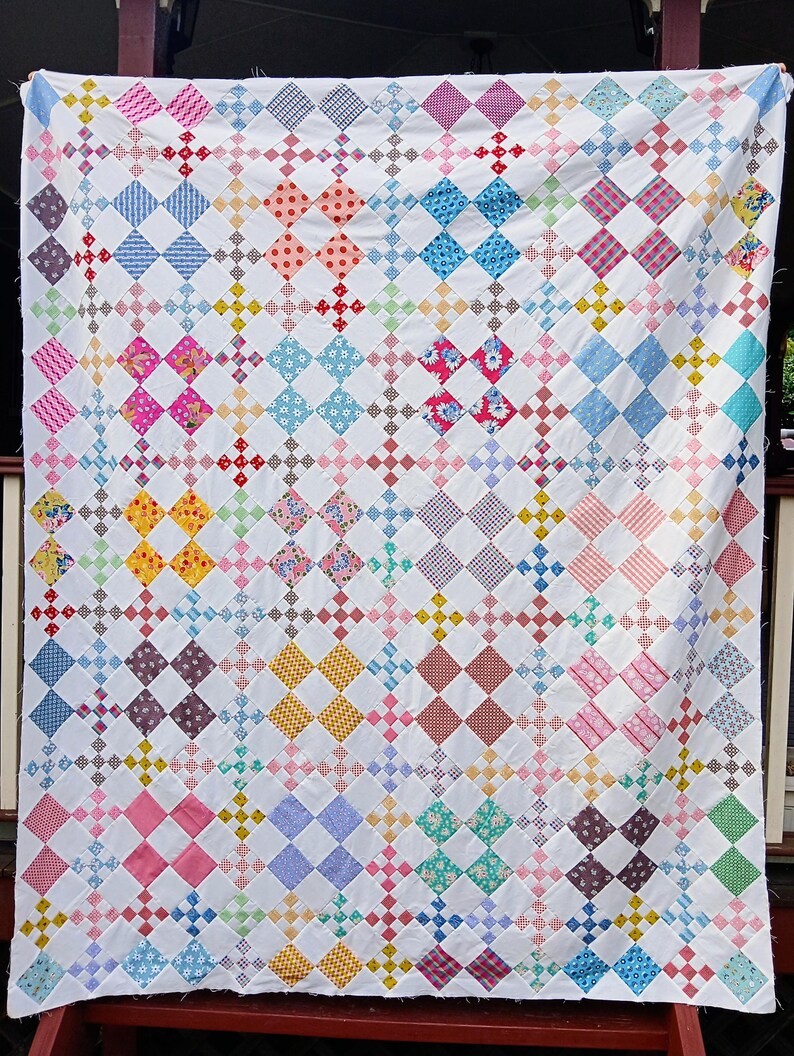 May include: A handmade quilt with a white background and a repeating pattern of colorful diamond shapes. The diamonds are filled with various patterns and colors, including blue, pink, yellow, and red. The quilt is displayed outdoors.