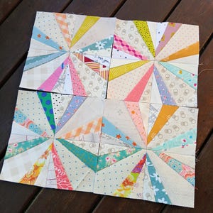 May include: Eight colorful quilt blocks arranged on a wooden surface. Each block features a sunburst design with various patterned fabrics in shades of pink, blue, yellow, and green. The blocks are approximately 8 inches square.