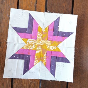 May include: A square quilt block with a star design. The star is made from pink, purple, and yellow floral fabric. The background is white with small dotted patterns and cross stitch details. The block is on a wooden surface.