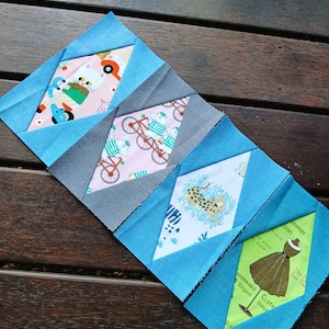 Puede incluir: Cuatro cuadrados de tela cosidos juntos en forma de diamante. Cada cuadrado presenta un estampado colorido diferente, que incluye un gato montando en bicicleta, una bicicleta con un ciclista, un guepardo y un vestido.