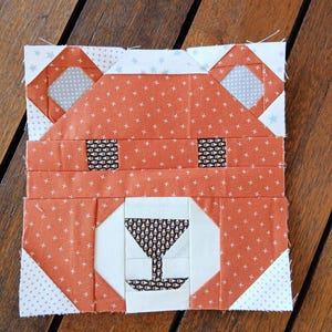 May include: A patchwork bear face made of orange fabric with white cross patterns. The bear has patterned eyes and a white nose with a patterned design. The ears are white with gray diamond shapes. The background is a wooden surface.