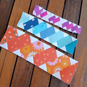 May include: Four colorful fabric bookmarks with a chevron design are displayed on a wooden surface. The bookmarks feature various patterns, including floral, geometric, and polka dots, in shades of orange, pink, blue, and purple. Each bookmark is approximately 1 inch wide.