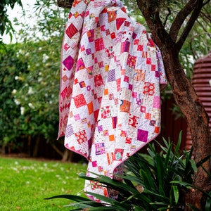 Puede incluir: Una colcha blanca con cuadrados rosas, rojos y naranjas. La colcha está colgada de una rama de árbol.