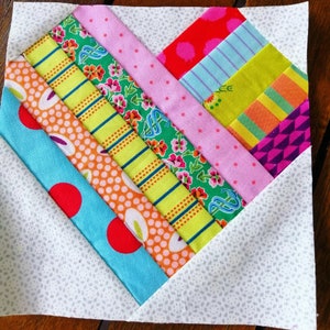 May include: A patchwork quilt block featuring a heart shape design. The heart is constructed from strips of fabric in a variety of colours and patterns, including pink, green, yellow, orange, blue, and red. The background fabric is white with a subtle speckled pattern.