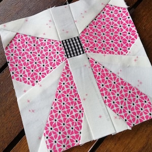 May include: A white fabric square with a pink and black floral pattern in the center. The fabric square is divided into eight sections with a white fabric border.