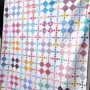 May include: A handmade quilt with a white background and a repeating pattern of colorful diamond shapes. The diamonds are filled with various patterns and colors, including pink, blue, yellow, and red. The quilt is hanging, showing its full size.