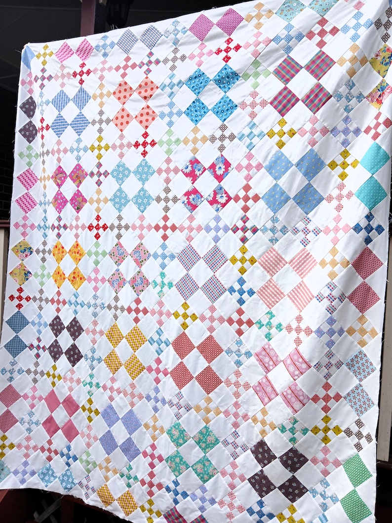 May include: A handmade quilt with a white background and a repeating pattern of colorful diamond shapes. The diamonds are filled with various patterns and colors, including pink, blue, yellow, and red. The quilt is hanging, showing its full size.