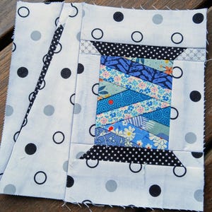 Op de afbeelding: Een stoffen quiltblok met een spoelontwerp. De spoel is gemaakt van verschillende blauwe stoffen met patronen. De achtergrond is wit met zwarte en grijze stippen. Het blok heeft een vierkante vorm.