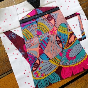 May include: A patchwork quilt block featuring a colorful floral pattern with a teal, pink, and yellow color scheme. The block is surrounded by white fabric with small red squares.