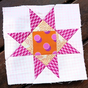 Può includere: Un blocco di quilt fatto a mano con un motivo a stella. La stella è composta da tessuto a fantasia rosa e bianco, con un centro arancione decorato con pois viola. Il blocco è quadrato, che mostra un'estetica patchwork.