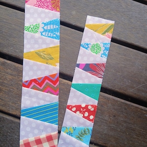 May include: Two handmade fabric bookmarks. Each bookmark features a light purple background with white polka dots. Colorful fabric triangles in pink, blue, yellow, and red are sewn onto the background, creating a patchwork design. The bookmarks are displayed on a wooden surface.